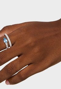 Anello d'argento con una fascia testurizzata che presenta un'ambientazione con una gemma blu a forma di pera, indossato su una mano dalla pelle scura su uno sfondo bianco.