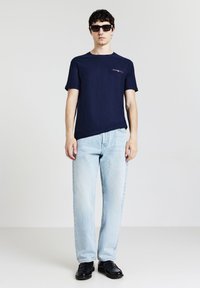 Uomo con occhiali da sole neri, T-shirt blu navy con piccolo taschino, jeans azzurri e scarpe nere, in piedi contro uno sfondo neutro.