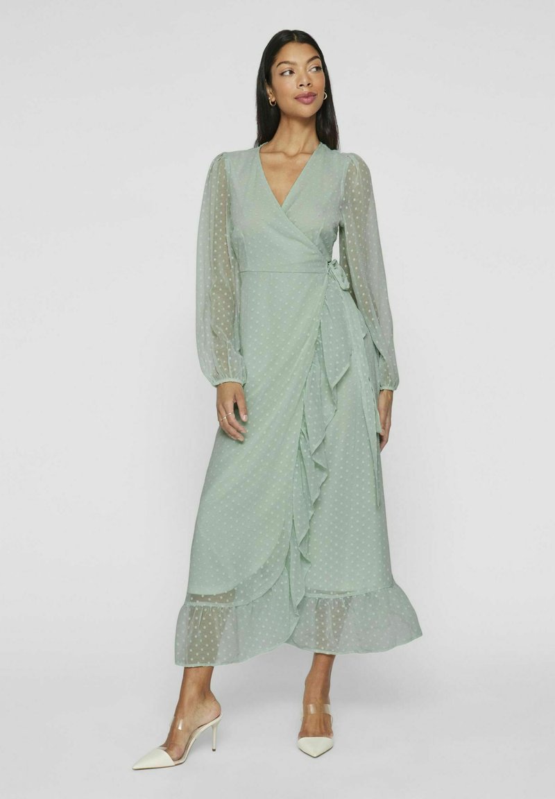 Mintgrønn, prikkete omslagskjole med lange, gjennomsiktige ermer, volangdetalj og knyttet midje. Glatt tekstur og ankel-lengde hem.