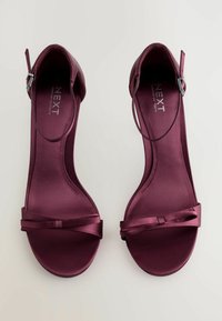 Sandalias de satén burdeos con tacón y una cinta delgada con detalle de lazo en la parte delantera del pie. Presenta una correa trasera con una hebilla de metal para el cierre.