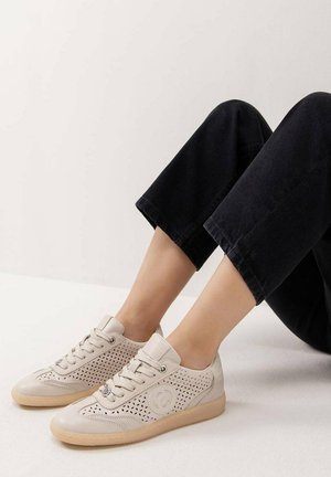 Persona que lleva zapatillas beige claro con cordones, patrón perforado y suelas beige, vestida con pantalones negros recortados, sentada en un suelo blanco.