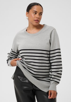 Femme portant un pull gris à rayures horizontales noires et un pantalon en cuir noir, tenant l'ourlet du pull d'une main.