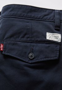 Pantalon bleu marine foncé en tissu tissé, avec fermeture à bouton, poche arrière avec étiquette et détail de surpiqûres rouges.