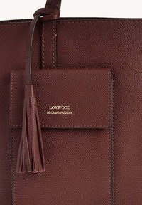 Sac fourre-tout en cuir texturé marron avec une étiquette en relief doré, comprenant une poche amovible et un détail à frange sur la fermeture éclair.