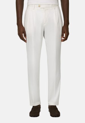 Pantalon blanc pour homme, coupé sur mesure, avec plis, poches latérales, passants pour ceinture, et mocassins en daim marron sur un fond uni.