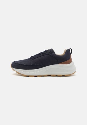 Sapatilha de camurça azul marinho com sola de borracha branca e bege, biqueira redonda e acabamentos texturizados. Apresenta um design com atacadores e uma aba traseira para facilitar o calçar.