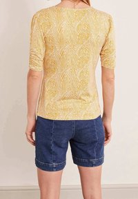 Kratký rukáv v žluté barvě s vzorem listů, s nařasením na rukávech. Kombinováno s modrými džínovými šortkami s viditelným ohrnutým lemem.