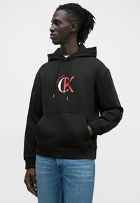 Sweatshirt preta com capuz e bolso frontal, com o logótipo Calvin Klein em vermelho e branco no peito. Confeccionada em tecido macio com punhos canelados.
