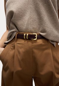 Brun läderbälte med guldfärgade detaljer, bärs över en beige stickad topp och bruna veckade byxor. Fokus på bältets textur och metall detaljer.