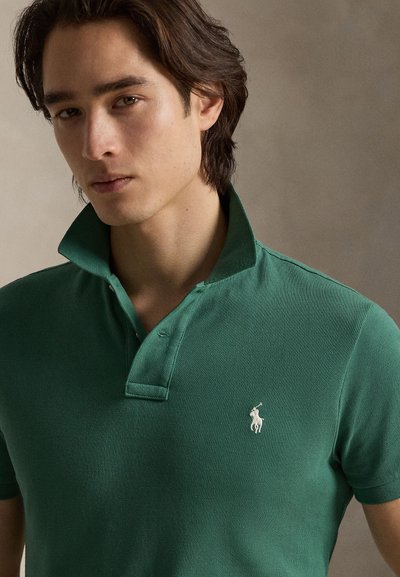 Polo verde feito de tecido texturizado, com uma pala de duas botões e um logótipo branco bordado no lado esquerdo do peito. A gola está levantada.