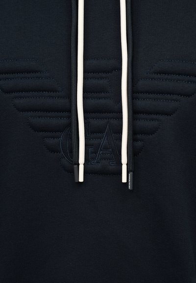 Μαύρο φούτερ με κουκούλα και ανάγλυφο ραμμένο λογότυπο με φτερά και γράμματα «GA», με αντίθετα μαύρα και λευκά κορδόνια με μπρελόκ με το εμπορικό σήμα.