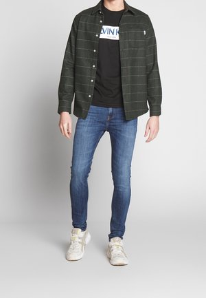 Camisa de franela a rayas verdes sobre una camiseta negra, combinada con jeans azules y zapatillas blancas. Presenta un cierre frontal de botones y un bolsillo lateral.
