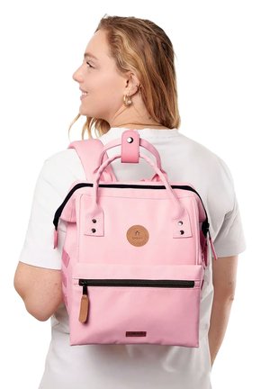 Person trägt weißes Hemd und trägt einen hellrosa Rucksack mit schwarzem Reißverschluss und beiger Leder-Logoplakette auf dem Rücken.