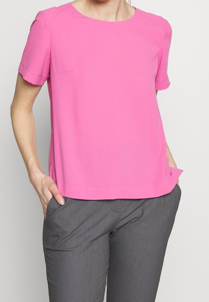 Femme portant un chemisier rose vif à manches courtes et un pantalon gris à carreaux avec les mains dans les poches, sur un fond uni.