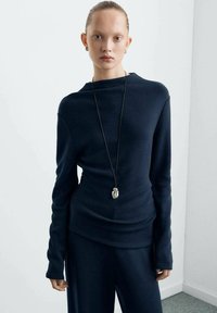 Top a manica lunga blu navy con un leggero scollo a barca e silhouette aderente, abbinato a pantaloni scuri coordinati. Collana con ciondolo d'argento che valorizza il look.