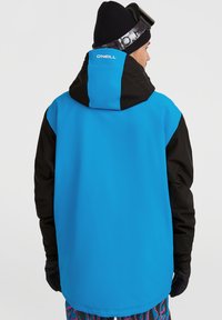 Chaqueta de snowboard azul y negra con capucha, que presenta una textura suave, un diseño casual y un logo en la parte posterior. Se llevan guantes negros.