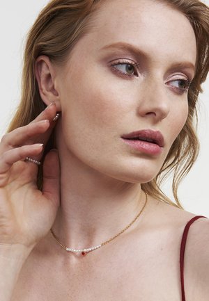 Guldhalsband med vita pärlor och en röd hängsmycke. Modellen bär pärlbeströdda örhängen, med avslappnat hår och neutral makeup.