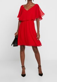 Robe en chiffon rouge avec un décolleté en V, des manches courtes volantées et une taille élastique. Présente un ourlet au genou et est associée à des accessoires noirs.