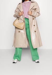Beige glänsande trenchcoat med stora knappar, kombinerad med en ljuslila skjorta, gröna byxor och en liten beige axelväska. Vita sneakers med gröna accenter.