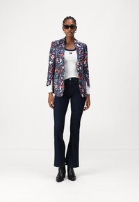 Blazer azul marino con estampado floral y mangas largas, combinado con una camiseta sin mangas blanca y jeans de campana oscuros. La modelo lleva botas negras hasta el tobillo y gafas de sol.