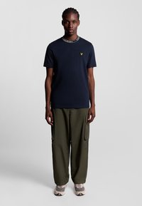 Mørkeblå kortærmet t-shirt med en hvid logo-kant, parret med olivengrønne cargo-bukser og beige sneakers. Rent design, afslappet pasform.