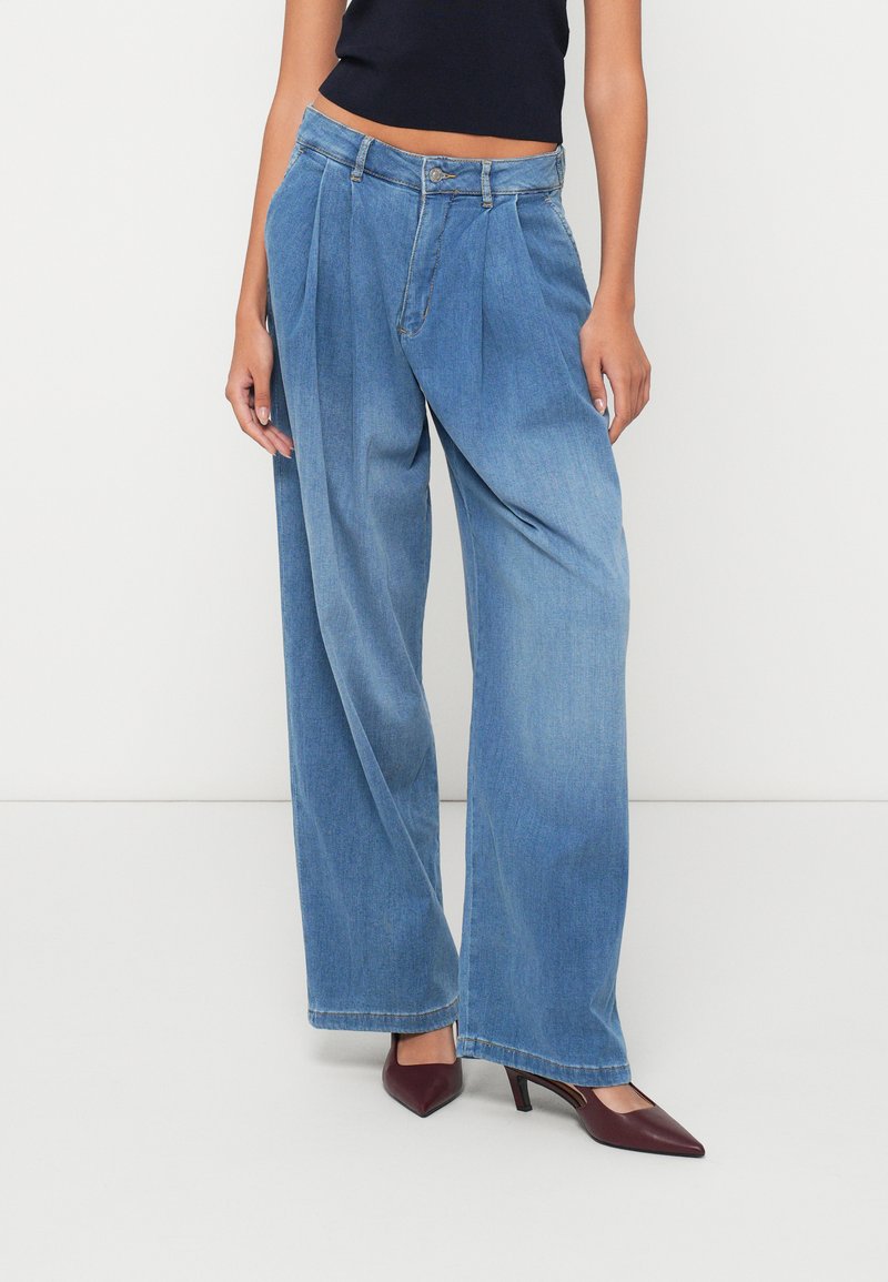 Jeans de pierna ancha azul claro con pliegues, hechos de denim. Presenta un cierre de botón y un ligero desvanecimiento, combinados con tacones puntiagudos burdeos.