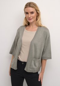 Šedý, kovový pletený kardigan s krátkými rukávy, otevřeným předním dílem a dvěma předními kapsami, zkombinovaný se světle béžovým topem a tmavými kalhotami.