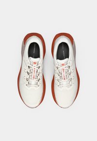 Miesten juoksukengät valkoisesta verkkomateriaalista, joissa punaiset kumikoristeet. Niissä on teksturoitu päällinen, pehmustettu kaulus ja heijastavat kengännauhat. Kevyt muotoilu.