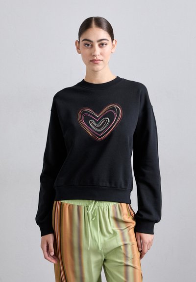 Femme portant un sweat à capuche noir avec un design de cœurs concentriques multicolores et un pantalon vert clair avec des rayures verticales orange et marron.