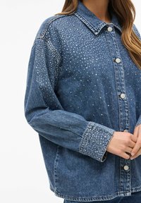 Jean jakna u svijetloplavoj boji, oversized kroja, ukrašena s rhinestones na prednjoj strani i rukavima. Srebrni zakovice.