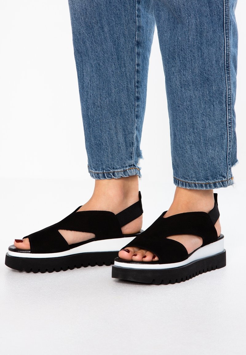 Sandales en suède noir à bouts ouverts, avec une semelle épaisse blanche et une semelle extérieure noire antidérapante. Le design comprend des découpes décoratives et une bride autour de la cheville.