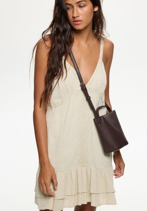 Beige jurk met V-hals, kantdetails en een gerimpelde zoom, gecombineerd met een kleine bruine tas van textuur met een schouderband.