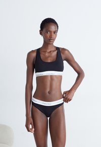 Sutiã desportivo preto com faixa logo branca da Calvin Klein, parte de baixo de bikini preto combinando com logo. Design simples, forma ajustada, textura de tecido suave.