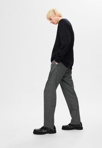 Schwarzer gestrickter Pullover, graue karierte Hose, schwarze Lederschuhe mit strukturierten Details, im Profil auf einem schlichten Hintergrund stehend.