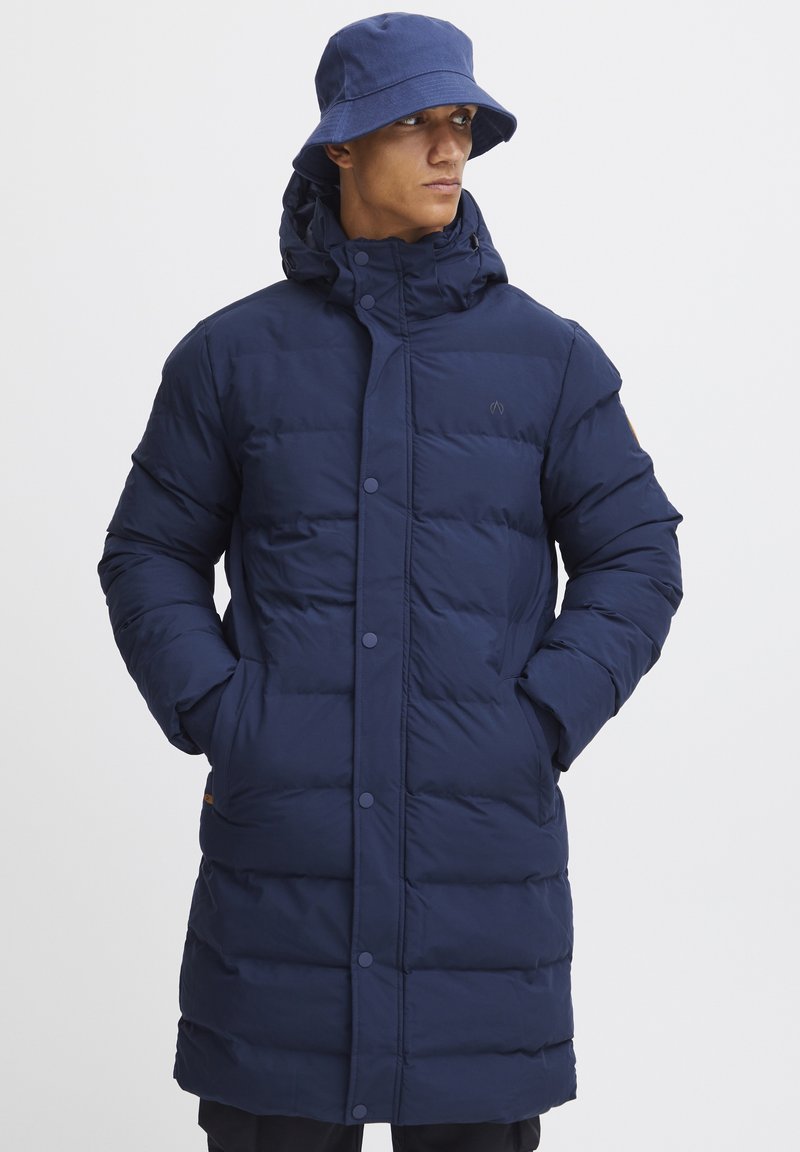 Navy blå puffjakke med hette, med store rektangulære paneler, trykknapper og to sidelommer. Båret med en matchende bøttehatt.