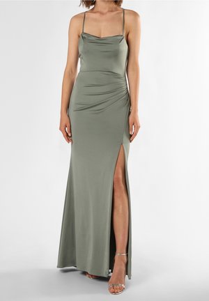Žena v dlouhých olivově zelených saténových šatech s tenkými ramínky a vysokým rozparkem, doplněných stříbrnými sandály s odkrytými prsty.