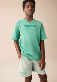 Grøn bomulds T-shirt med "SUB5CRIBE" i en mørkere grøn farve, matchende med lyse grønne shorts, der har den samme tekst. Simpelt design, korte ærmer.