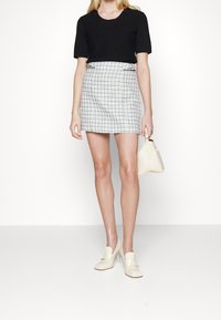 Mujer con blusa negra de manga corta, falda corta de cuadros blancos, zapatos de tacón color crema y sosteniendo un bolso color crema.