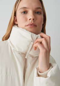 Giacca puffer bianca con colletto alto, dotata di chiusura a zip e foderatura morbida e texturizzata. Il tessuto appare liscio e lucido.