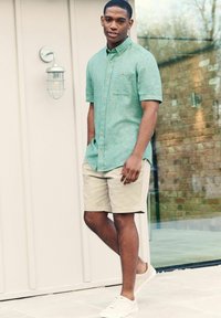 Ljusgrön kortärmad skjorta med knappar, beige shorts och vita sneakers som bärs av en manlig modell stående utomhus nära en glasvägg och en ljuskälla.