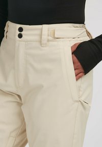 Beige broeken met twee zwarte knopen op de taille, een zijzak en een merklabel. Fijne textuur met een casual pasvorm.