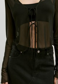Schwarzes transparentes Langarmoberteil mit einem cropped Design, ausgestattet mit einem vorderen Bindedetail, kombiniert mit einem enganliegenden schwarzen Jeansrock.