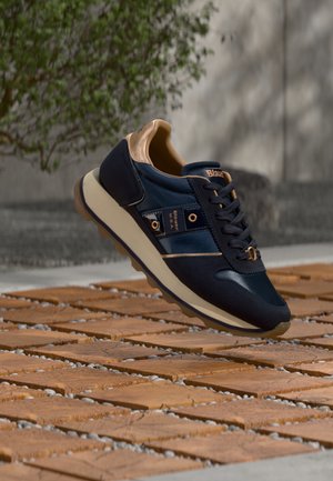 Scarpa da ginnastica blu navy e beige con suola spessa e lacci, etichettata "Blauer U.S.A.", che fluttua sopra un blocco di legno e un terreno con motivo di ciottoli all'aperto.