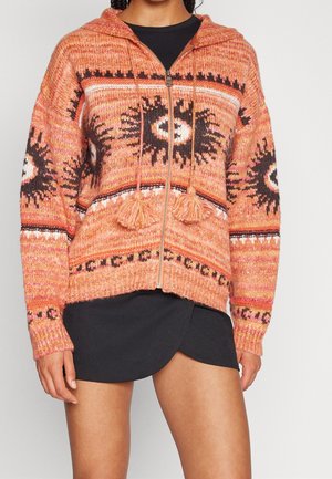 Femme portant un pull à capuche orange zippé avec des motifs rayons de soleil noirs et blancs et des pompons, associé à une jupe portefeuille noire.