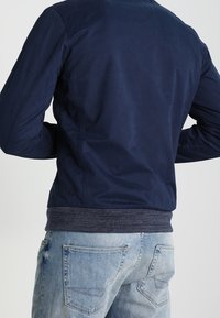 Marineblauwe bomberjack van glad materiaal, met geribbelde manchetten en zoom, voorzien van een subtiel logodetail op de achterkant. Gecombineerd met lichtblauwe spijkerbroek.