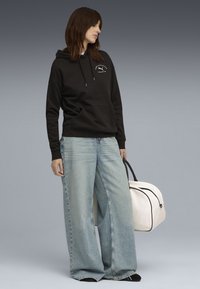 Sort hættetrøje med frontlomme, parret med brede lyseblå jeans. En hvid weekendtaske med sorte detaljer bæres i den ene hånd.