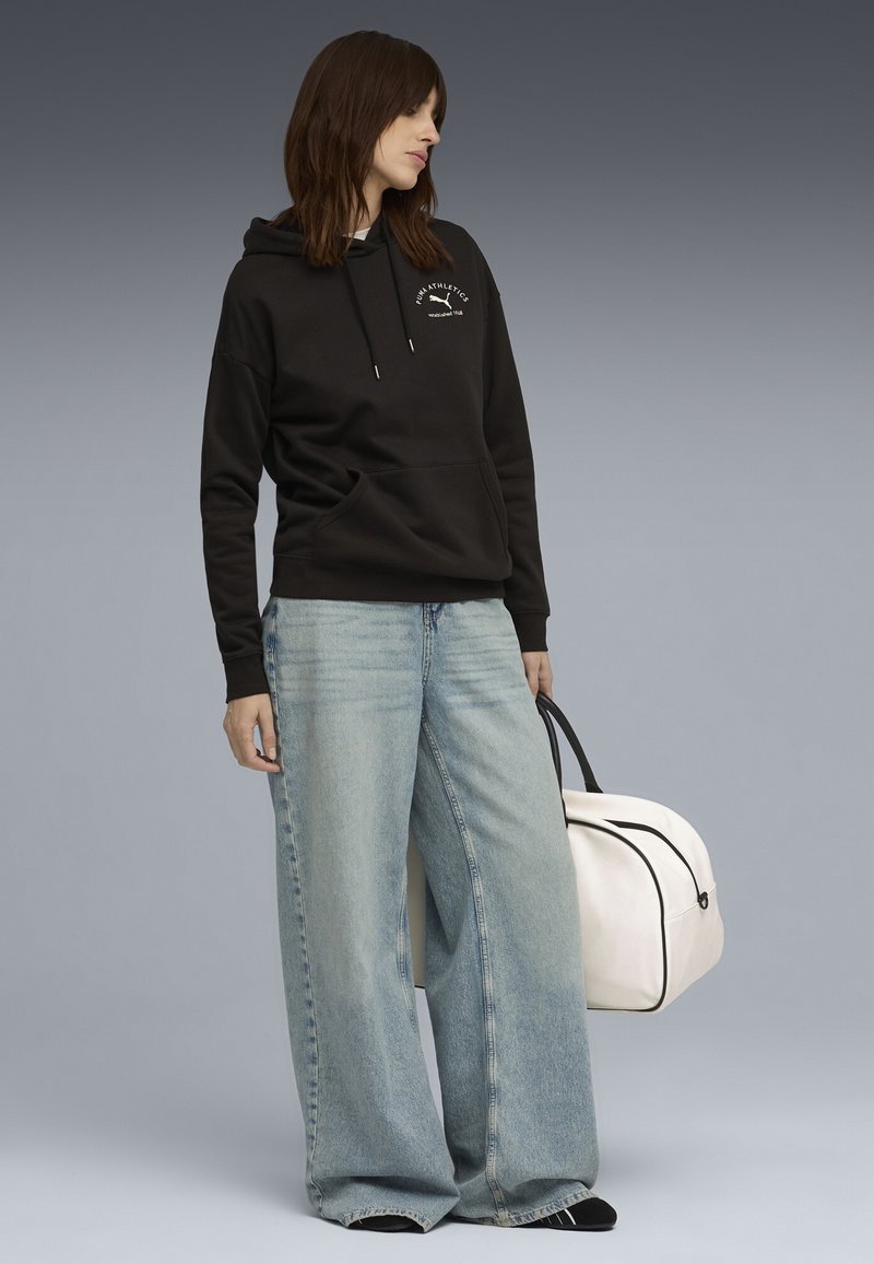 Sort hættetrøje med frontlomme, parret med brede lyseblå jeans. En hvid weekendtaske med sorte detaljer bæres i den ene hånd.