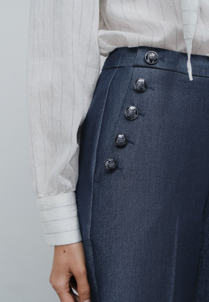 Gros plan d'une personne portant un pantalon bleu foncé taille haute avec cinq boutons métalliques décoratifs sur le côté et une chemise rayée de couleur claire.
