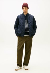 Marineblauwe gequiltte bomberjack, gestreepte polo, olijfgroene broek en witte sneakers met bruine zolen; kastanjebruine gebreide beanie. Eenvoudig, strak design.