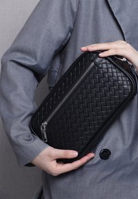 Black woven leather bag with a silver zipper and sleek edges. The bag is held against a gray fabric background, enhancing its texture.